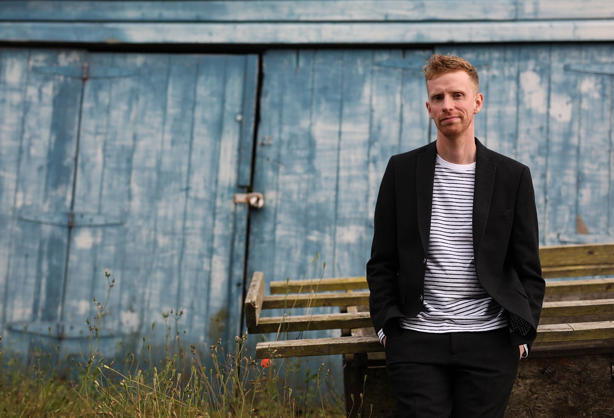 An image of singer-songwriter Adam Ross standing outdoors. He is wearing a black blazer over a striped t-shirt and black trousers. He is standing in front of two large, blue barn doors.