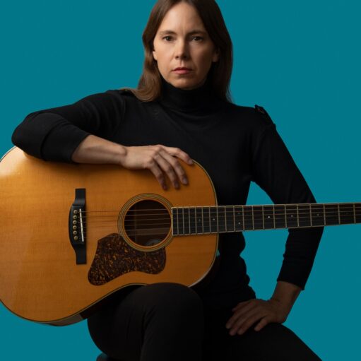 An image of singer-songwriter Bronwynne Brent sitting on a stool and holding an acoustic guitar. She's wearing a black long-sleeved turtleneck and black trousers and is looking directly at the camera. The background is solid turquoise.