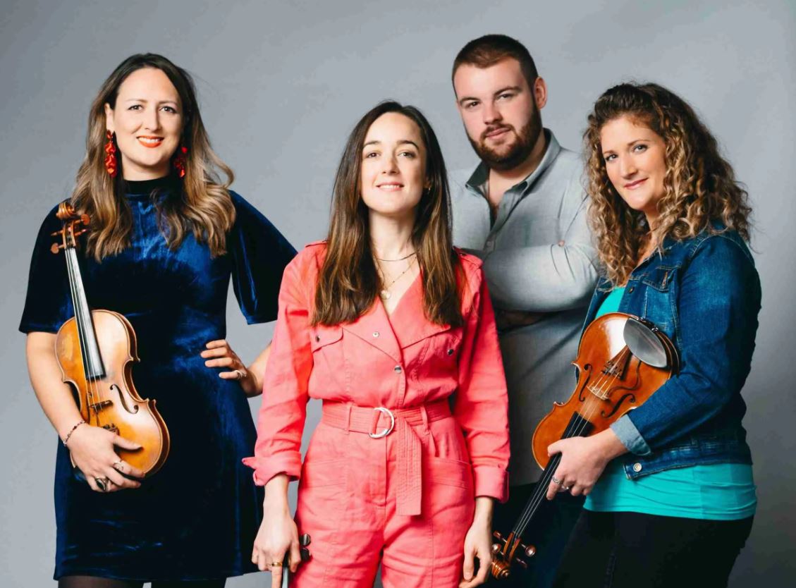 A studio portrait of four musicians standing in front of a gray background. From left to right: a woman in a dark blue dress holding a violin; a woman in a pink jumpsuit; a man in a gray shirt with his arms crossed; and a woman in a denim jacket holding a violin. They are all looking at the camera with confident, relaxed expressions.