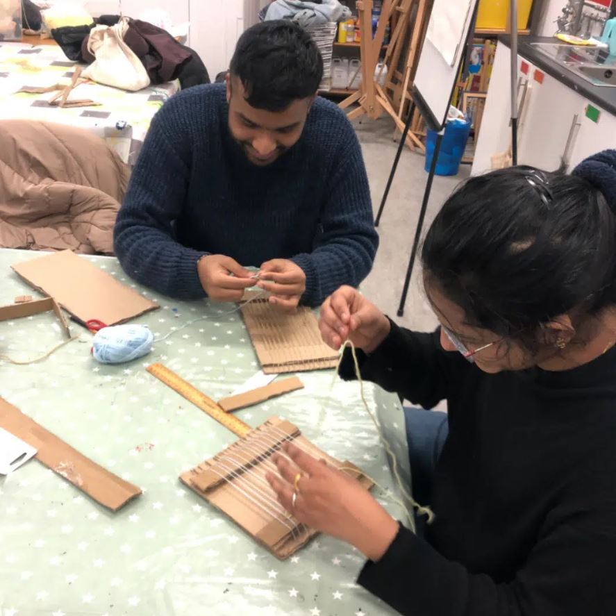 Two people are seated at a table, weaving with yarn on cardboard looms. The person on the left is wearing a blue sweater and smiling down at their work. The person on the right, wearing a black top and glasses, is focused on their weaving project. On the table are pieces of cardboard, a ruler, and a ball of light blue yarn.