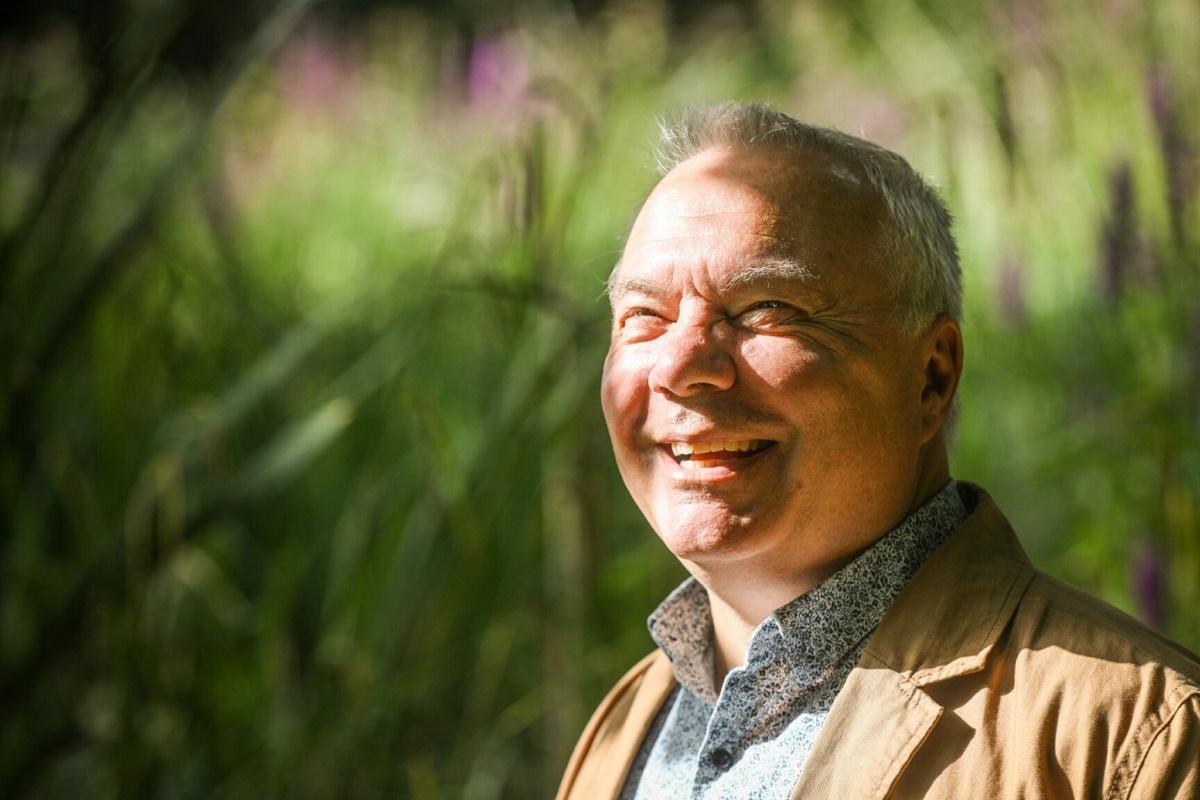 A close-up outdoor portrait of a smiling, fair-skinned man with short grey hair, looking slightly up and to the left. He is wearing a light brown jacket over a patterned collared shirt. Bright sunlight illuminates the right side of his face. The background is a soft, out-of-focus blend of bright green and hints of purple and yellow foliage.