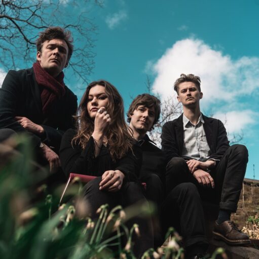 An image of the band Kirsten Adamson and the Tanagers sitting outdoors. There are four people in the image. Two men are on the left side, and one man and one woman are on the right side. The person on the far left and the two people on the right are looking into the camera. The person in the front on the left is looking away from the camera. The sky is bright blue and there are yellow daffodils in the foreground.