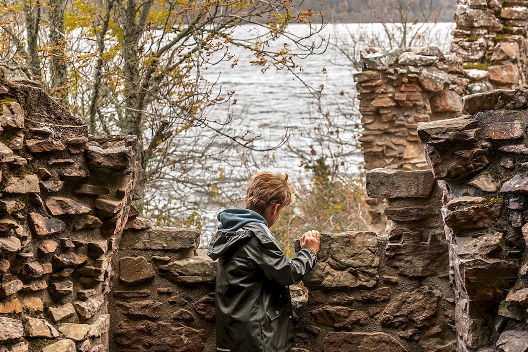A child in a green jacket and blue hoodie is looking out from behind a stone wall. The stones are part of a ruined structure. Beyond the wall, a large body of water is visible, with trees on the far shore.