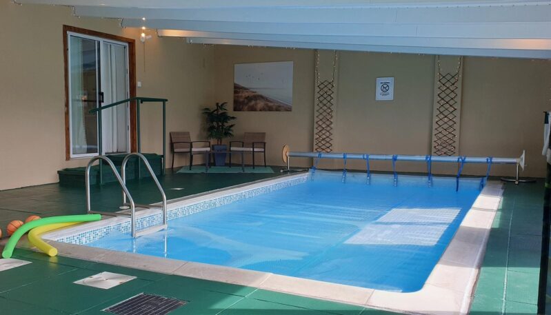 An indoor swimming pool with a blue cover pulled over the water. The walls are a light tan color, and the floor is a dark green tile. There are some pool noodles and balls on the left side of the pool, and a small table with chairs is visible on the far side.