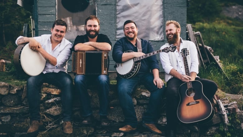 An image of the band Rum Ragged. The four band members are sitting on a stone wall in front of a weathered-looking green building. The man on the left is holding a bodhrán, the second man from the left is holding an accordion, the man to his right is holding a banjo, and the man on the far right is holding a guitar. All four are smiling at the camera.