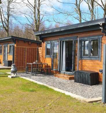 Two rustic, wooden cabins with dark gray trim and large windows sit side-by-side in a grassy, outdoor area. Each cabin has a small patio with a table and chairs, surrounded by light-colored gravel. Trees and a small fence are visible in the background under a blue sky.