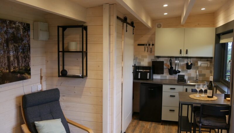 A modern, compact kitchen inside a wood-paneled cabin. The kitchen has light green cabinets, a gray countertop, and a small dining table with two chairs. A mini-fridge and coffee maker are visible on the counter, and a framed picture of a forest hangs on the wall.