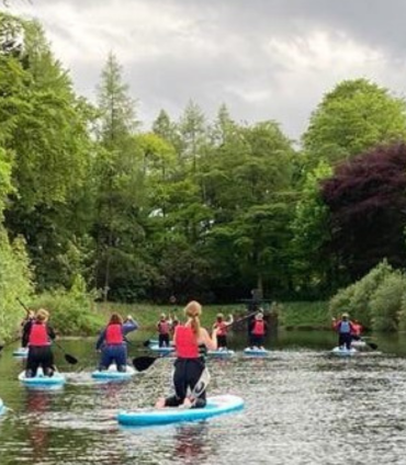 A group of people on paddleboards, mostly on their knees, are paddling down a calm river. They are all wearing red or dark life vests. Lush green trees and foliage line both banks of the river under a cloudy sky.